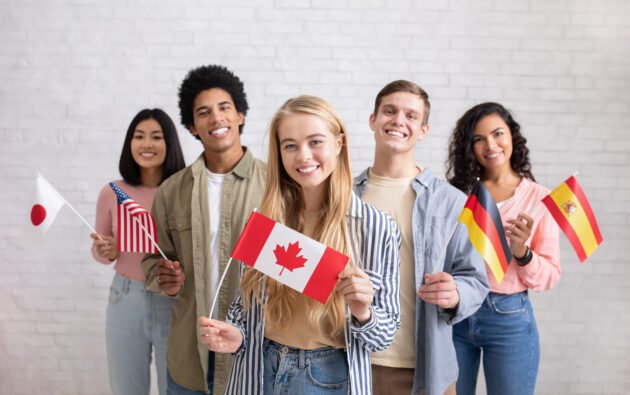 International exchange program and study together. Smiling young multi ethnic students looking at camera and holding flags of their countries, on white brick wall background, copy space, studio shot